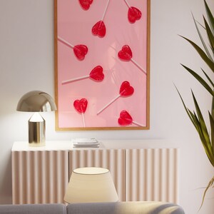 May include: A framed print of a pink background with multiple red heart-shaped lollipops on white sticks. The lollipops are arranged in a random pattern.