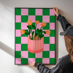 May include: A framed art print featuring a pink and green checkered background. A pink vase with orange flowers and green stems is centered on the print. The artwork is being held by a person.