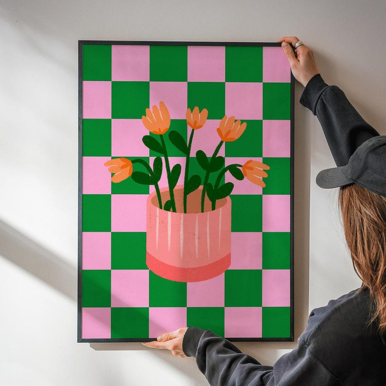 May include: A framed art print featuring a pink and green checkered background. A pink vase with orange flowers and green stems is centered on the print. The artwork is being held by a person.