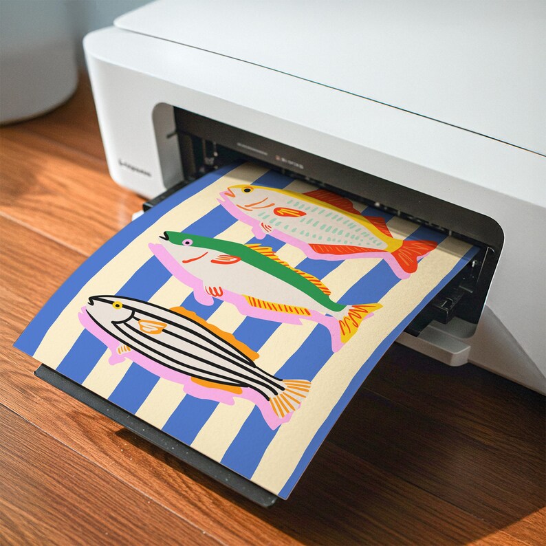 May include: A white printer is shown printing a colorful illustration of fish. The illustration features three fish with different patterns and colors, set against a blue and cream striped background. The fish have pink, green, yellow, and orange accents.