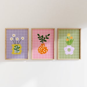 May include: Three framed art prints showcasing floral arrangements in vases. The prints have a grid pattern background in purple, pink, and green. The vases are yellow, orange, and lavender, with white daisies, green leaves, and orange flowers.