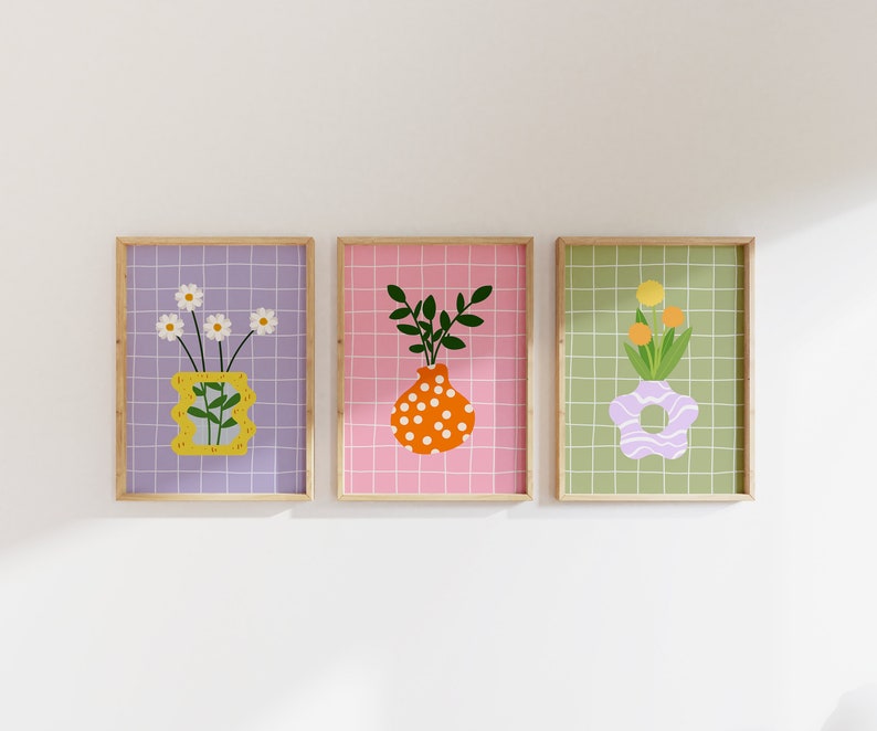 May include: Three framed art prints showcasing floral arrangements in vases. The prints have a grid pattern background in purple, pink, and green. The vases are yellow, orange, and lavender, with white daisies, green leaves, and orange flowers.