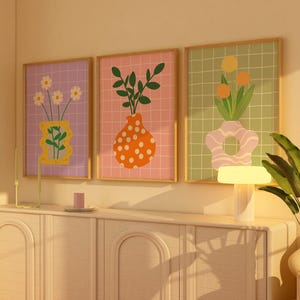 May include: Three framed art prints with floral illustrations in vases. The prints have a grid pattern background in purple, pink, and green. The vases are in yellow, orange, and pink. The frames are a light wood colour.