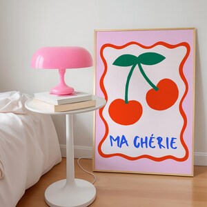 May include: A pink and gold framed print with a pink background. The print features two red cherries with green stems and leaves. The text "MA CH&Eacute;RIE" is written in blue below the cherries.