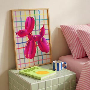 May include: A framed print featuring a pink balloon dog against a plaid background. A green book with the word "ROME" is on a green tiled surface, along with a blue and white striped mug. Pink striped pillows are in the background.