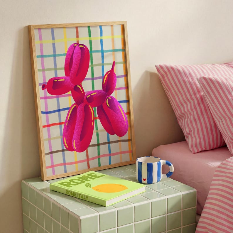 May include: A framed print featuring a pink balloon dog against a plaid background. A green book with the word "ROME" is on a green tiled surface, along with a blue and white striped mug. Pink striped pillows are in the background.