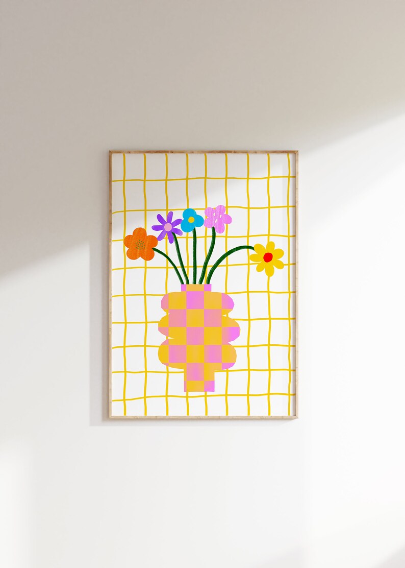 May include: A framed wall art print featuring a checkered pink and yellow vase holding five colorful flowers in orange, purple, blue, pink, and yellow. The background consists of a white surface with a hand-drawn yellow grid pattern throughout.