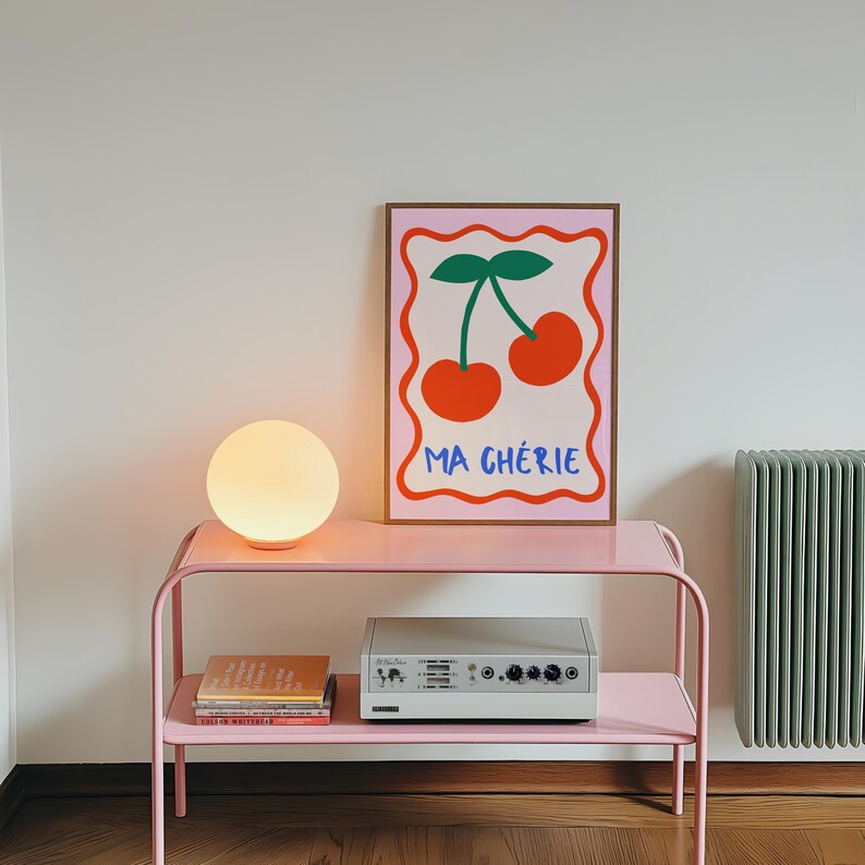 May include: A pink metal shelf with two shelves, a white globe lamp, a stack of books, and a white stereo system. A framed print with a pink background and a graphic of two red cherries with green stems and leaves. The print has the text "MA CH&Eacute;RIE" in blue and red letters.
