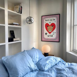 May include: A rectangular wall art print in a black frame featuring a red heart with a leopard face inside, surrounded by the text Born to be wild, small star icons, and a pink background with a green dashed border, displayed on a white wall.