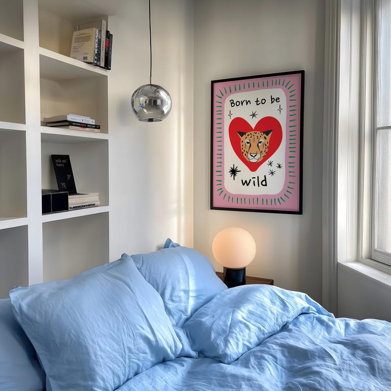 May include: A rectangular wall art print in a black frame featuring a red heart with a leopard face inside, surrounded by the text Born to be wild, small star icons, and a pink background with a green dashed border, displayed on a white wall.