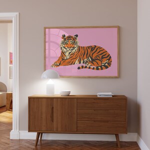 May include: A brown wooden dresser with three drawers and two doors. The dresser has a white lamp and a bowl on top. A framed print of a tiger lying down on a pink background is hanging on the wall above the dresser.