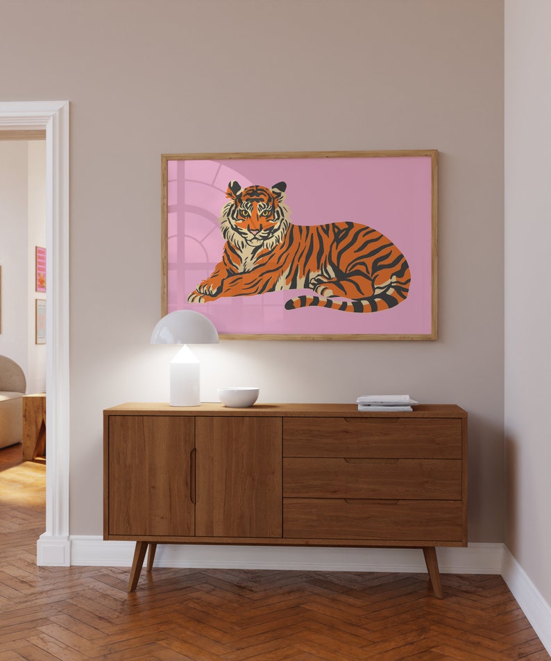 May include: A brown wooden dresser with three drawers and two doors. The dresser has a white lamp and a bowl on top. A framed print of a tiger lying down on a pink background is hanging on the wall above the dresser.