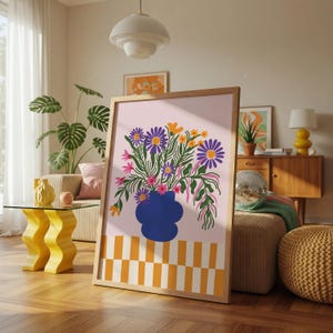 以下が含まれることがあります： 青い花瓶に活けられた鮮やかな花の絵がフレームに入っています。アートワークは、紫、黄色、ピンクの花を淡いピンクの背景に配置し、下部に市松模様があります。