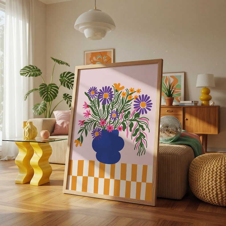 以下が含まれることがあります： 青い花瓶に活けられた鮮やかな花の絵がフレームに入っています。アートワークは、紫、黄色、ピンクの花を淡いピンクの背景に配置し、下部に市松模様があります。