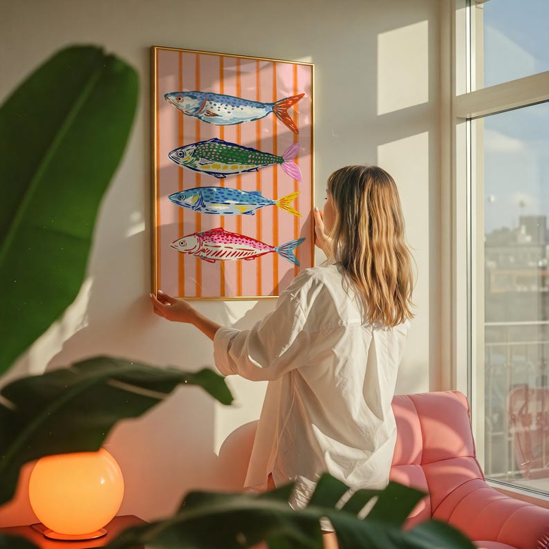 May include: A framed art print featuring four colorful fish illustrations against a pink and white striped background. The fish have blue, green, and red bodies with orange, pink, and yellow fins. The frame is gold.