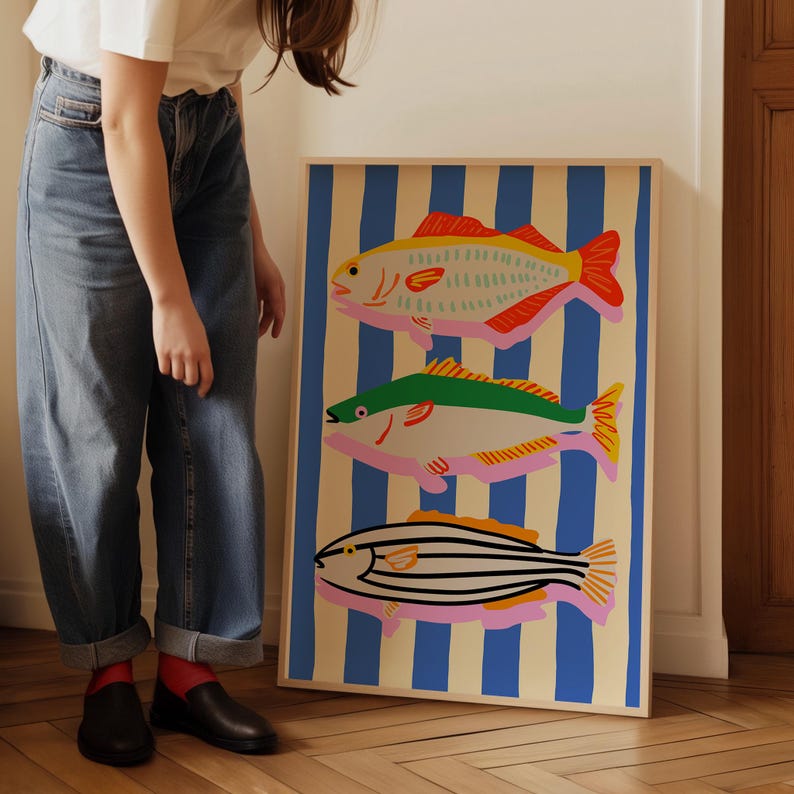 May include: A framed art print featuring three stylized fish illustrations. The fish are in shades of red, green, and black, with pink and yellow accents, set against a background of blue and cream vertical stripes. The artwork is displayed indoors.