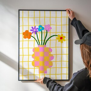May include: A framed wall art print featuring a checkered pink and yellow vase holding five colorful flowers with green stems. The background consists of a yellow grid pattern on a white surface. The flowers are orange, purple, blue, pink, and yellow.