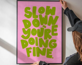 May include: A framed pink poster with the words "SLOW DOWN YOU'RE DOING FINE" in large, green, bubble letters. The poster is held by a person wearing a black cap and a black long-sleeved shirt.