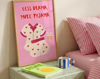 May include: A framed pink print with the text "LESS DRAMA, MORE PYJAMA." The print features a pajama set illustration with red hearts, and two pairs of slippers. A blue and white striped mug and a book are on a green tiled surface.