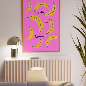 Może przedstawiać: Wydruk w ramie z r&oacute;żowym tłem i ż&oacute;łtymi bananami. Tekst "life is bananas" jest napisany na wydruku czarnym kolorem.