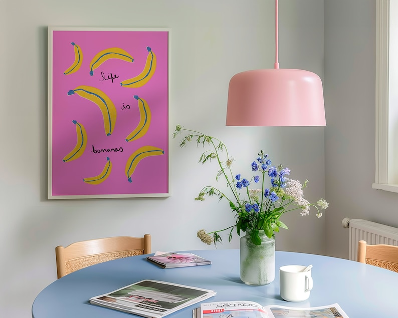 Może przedstawiać: R&oacute;żowo-biały wydruk w ramie z r&oacute;żowym tłem i ż&oacute;łtymi bananami z tekstem "life is bananas" w kolorze niebieskim. R&oacute;żowe wiszące światło znajduje się nad niebieskim stołem z wazą z niebieskimi i białymi kwiatami oraz białym kubkiem.