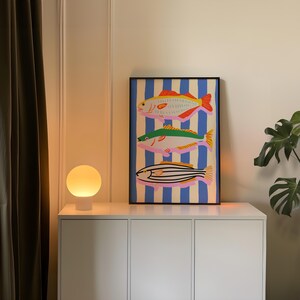 May include: A framed art print featuring three colorful fish illustrations against a blue and white striped background. The fish are in shades of yellow, green, and pink, with black outlines. The artwork is displayed on a white cabinet, next to a round, white lamp.