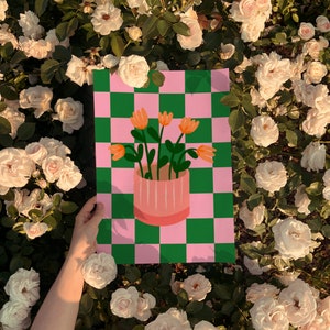 May include: A colorful art print featuring a pink and green checkered background. The print showcases a pink vase with orange flowers. The artwork is held against a backdrop of white roses.