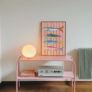 May include: A pink metal side table with a spherical lamp and a framed print of fish. The print shows four fish in blue, green, and red against a pink striped backdrop. A silver audio component rests on the lower shelf. The lamp emits a warm glow.