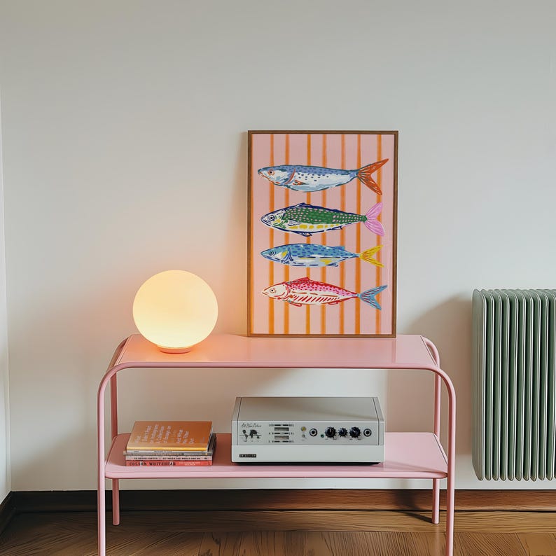 May include: A pink metal side table with a spherical lamp and a framed print of fish. The print shows four fish in blue, green, and red against a pink striped backdrop. A silver audio component rests on the lower shelf. The lamp emits a warm glow.