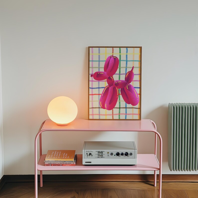 May include: A pink metal side table with two shelves holds a glowing orb lamp, books, and a silver audio device. A framed print of a pink balloon dog against a colorful plaid background hangs on the wall. A green radiator is visible.