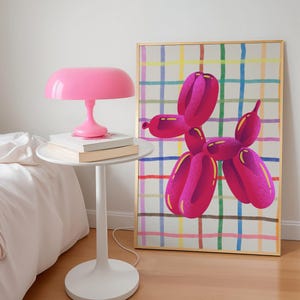 May include: A pink table lamp sits on a white side table with books. A framed artwork of a pink balloon dog with yellow accents hangs on the wall. The artwork has a gold frame and a colorful plaid background.