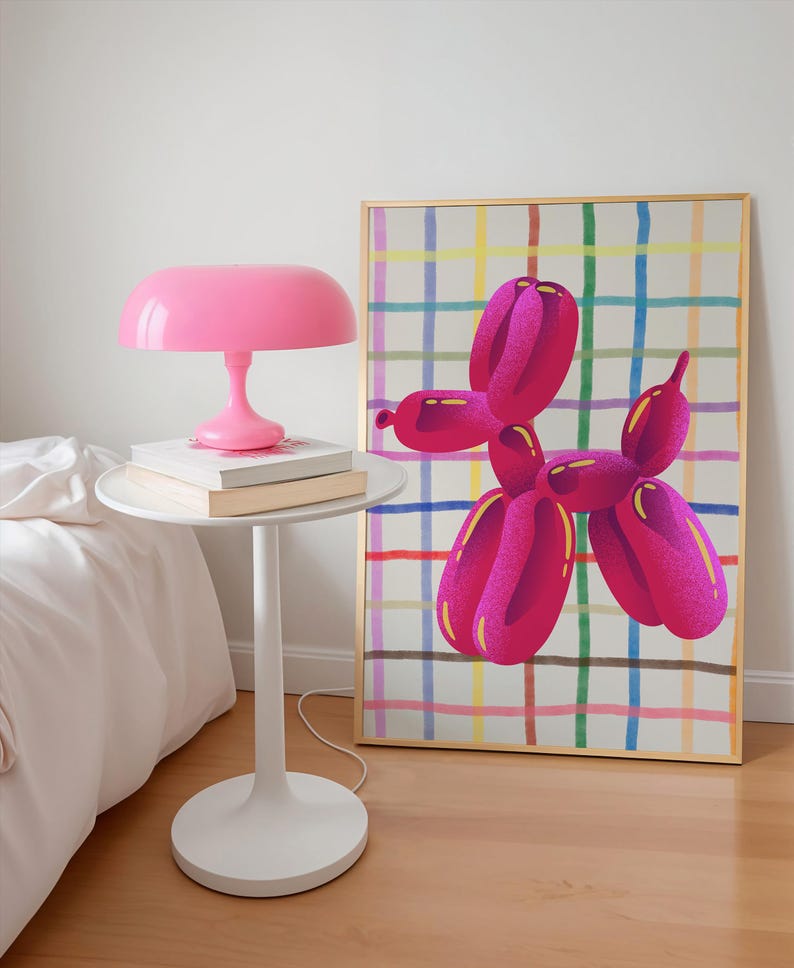 May include: A pink table lamp sits on a white side table with books. A framed artwork of a pink balloon dog with yellow accents hangs on the wall. The artwork has a gold frame and a colorful plaid background.