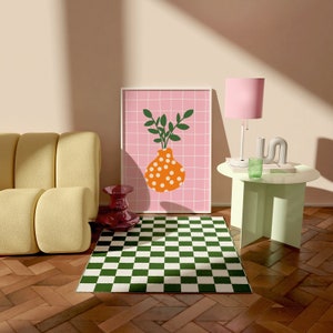 May include: An interior scene with a framed artwork depicting an orange vase with white spots and green leaves against a pink grid. A green and white chequered rug is on a wooden floor. A yellow sofa and a pink lamp are also present.