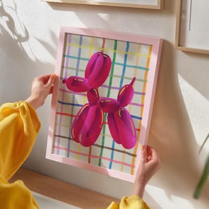May include: A pink framed print featuring a magenta balloon dog illustration. The dog is set against a colorful plaid background. The frame is a soft pink color. The artwork is displayed on a white wall.