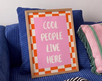 May include: A rectangular framed wall art piece featuring a pink center panel with the white text COOL PEOPLE LIVE HERE, surrounded by an orange and white checkered border. The print is encased in a light-toned wooden frame with a clean finish.