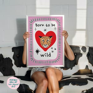May include: A rectangular wall art print in a silver frame featuring a white background with a large red heart containing a leopard face. The text Born to be wild is printed in black. The border is pink with green dashes and black star accents.
