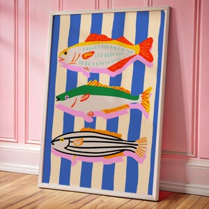 May include: A framed art print featuring three stylized fish illustrations. The fish are in shades of yellow, green, and black, with pink accents, set against a background of blue stripes and a cream color. The artwork is framed in a white border.