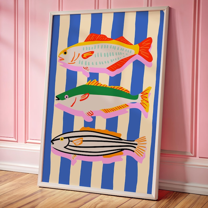 May include: A framed art print featuring three stylized fish illustrations. The fish are in shades of yellow, green, and black, with pink accents, set against a background of blue stripes and a cream color. The artwork is framed in a white border.
