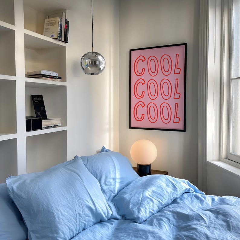 May include: A bedroom interior with a light blue bed set and pillows. A framed pink poster with the word "COOL" in red is on the wall. A chrome sphere pendant light hangs from the ceiling, and a round lamp sits on a bedside table.