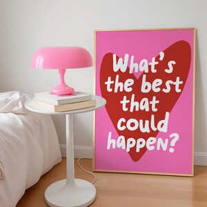 May include: A pink table lamp sits on a white side table with books. A framed pink poster with a red heart and the text "What's the best that could happen?" is propped against the wall. The room has a minimalist aesthetic.
