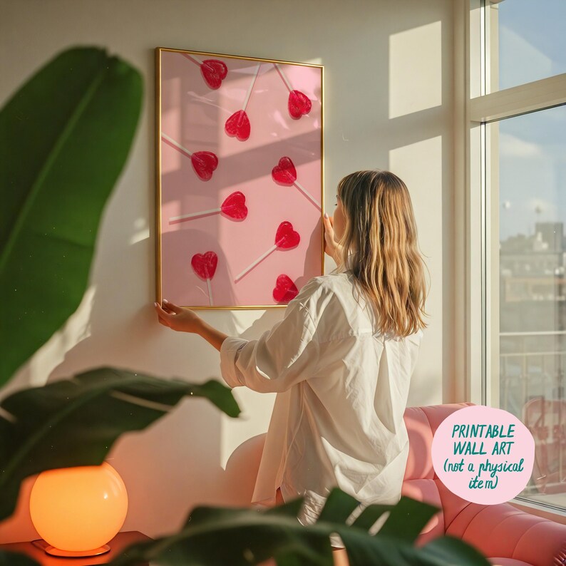 May include: A printable wall art print featuring a pink background with red heart-shaped lollipops on sticks. The print is framed in gold and is being hung on a white wall.