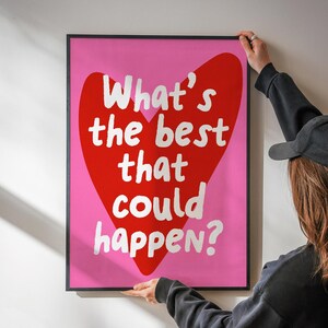 May include: A framed art print with a pink background and a large red heart shape. The text "What's the best that could happen?" is written in white inside the heart. The frame is black.