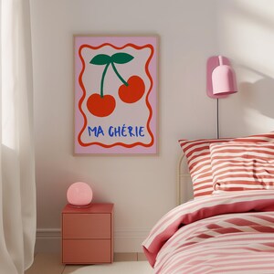 May include: A pink and white framed print with a red cherry illustration and the text "Ma Ch&eacute;rie" in blue and red letters. The print is hanging on a white wall above a red nightstand with a pink globe lamp.