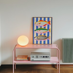 May include: A pink metal side table with a round, glowing lamp and a framed art print featuring three colorful fish against a blue and white striped background. A stereo and books are on the lower shelf. The wall is white.