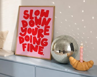 May include: A framed rectangular print with a light pink background and bold red text that reads SLOW DOWN YOU'RE DOING FINE. Next to it is a silver mirrored disco ball and a light pink spiral candle in a tan croissant-shaped candle holder.