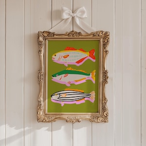 Puede incluir: Una impresi&oacute;n art&iacute;stica enmarcada con tres ilustraciones de peces coloridos sobre un fondo verde. Los peces son de color rosa, amarillo, rojo, verde y negro. El marco dorado ornamentado est&aacute; suspendido por una cinta blanca atada con un lazo.