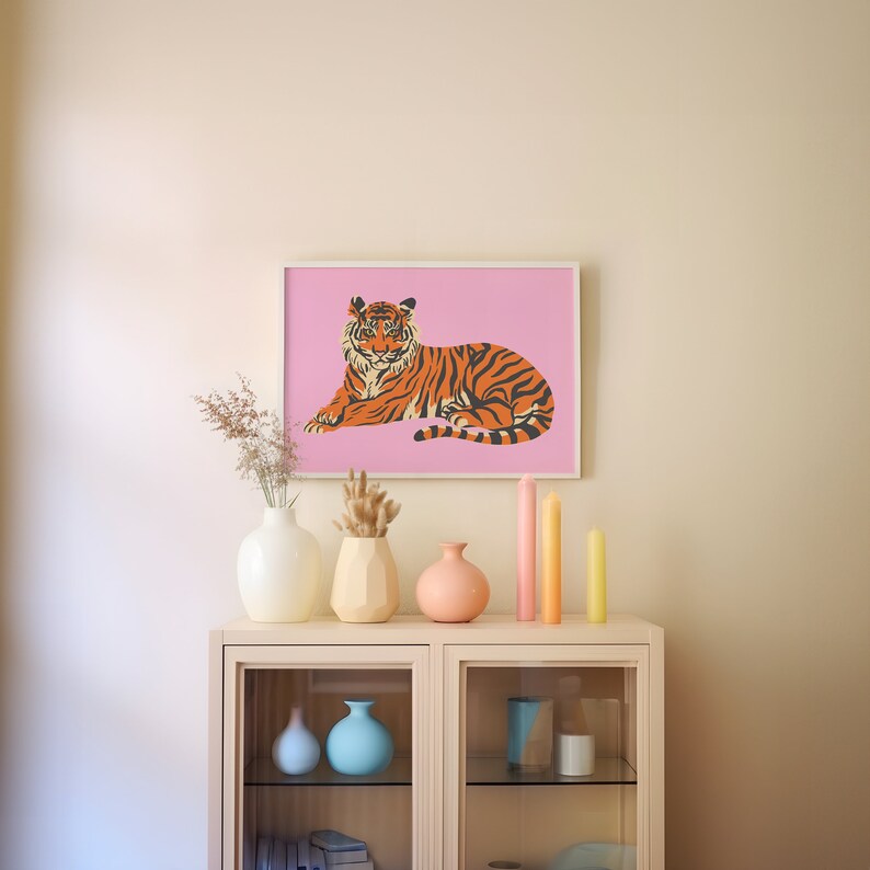 May include: A framed print of an orange and black tiger lying down on a pink background. The print is hanging on a light beige wall above a white shelf with vases and candles.