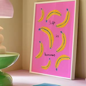 Może przedstawiać: Wydruk w ramie z r&oacute;żowym tłem i ż&oacute;łtymi bananami z niebieskimi konturami. Tekst "life is bananas" jest napisany czarnym kolorem na wydruku.