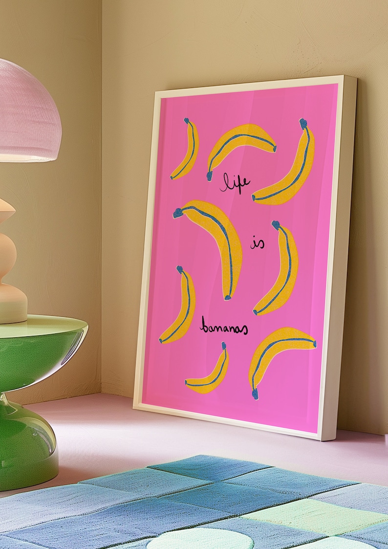 Może przedstawiać: Wydruk w ramie z r&oacute;żowym tłem i ż&oacute;łtymi bananami z niebieskimi konturami. Tekst "life is bananas" jest napisany czarnym kolorem na wydruku.