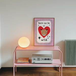 May include: A rectangular wall art print in a wooden frame featuring a leopard face inside a red heart on a pink background with green dashes around the border and the black text Born to be wild with small star icons surrounding the central heart.